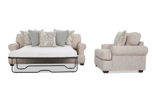 Beige fabric sofa bed with rolled arms and dark wooden legs, featuring a pull-out mattress and decorative paisley and solid gray cushions.