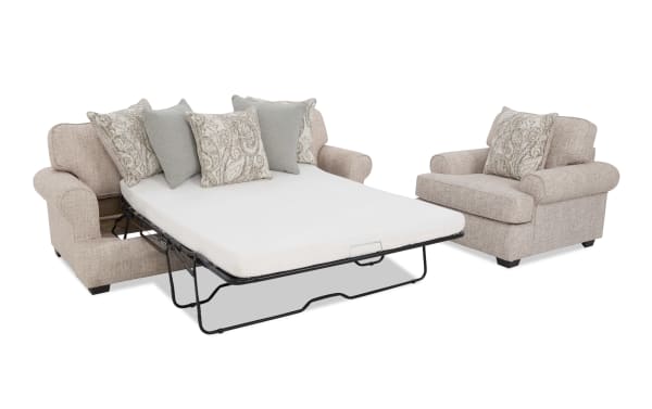 Beige fabric sofa bed with rolled arms, patterned and solid throw pillows, and a matching armchair, showcasing a pull-out mattress for guest sleeping.