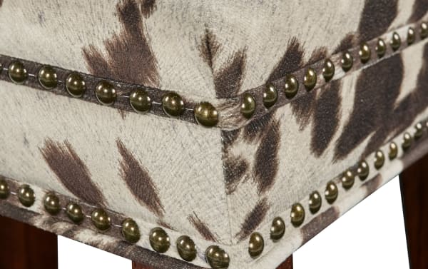 Close-up of Kristen Cow Print Walnut Backless Upholstered Counter Height Stool corner, showing brown and cream polyester fabric with cow print, walnut finish, and brass nailhead trim on traditional rubberwood frame.