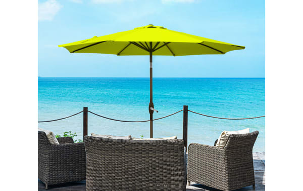 Kiley 9' lime green outdoor market umbrella with 360-degree tilt and crank system, polyester fabric, steel frame, and high-density plastic parts, shown with wicker patio furniture by the ocean.