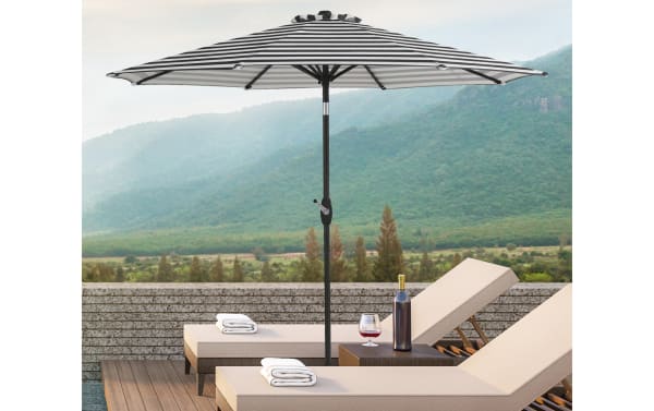 Kiley 9' black striped outdoor market umbrella with polyester fabric, steel pole, crank system, 360° rotation, and tilt, shown shading beige polyester lounge chairs.