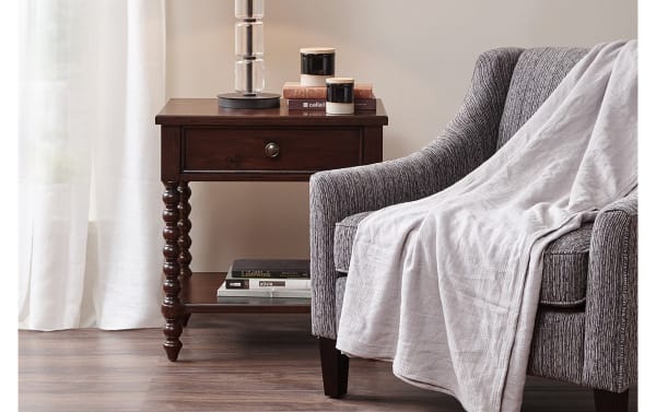 Traditional Padme Brown Nightstand with Drawer in acacia veneers, natural finish, turned wood legs, lower shelf, and warm farmhouse style next to gray armchair.