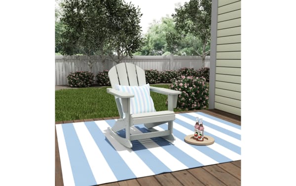 White Outdoor Sand Rocking Adirondack Chair in high-density plastic with transitional style, shown on blue and white striped outdoor rug with pillow.