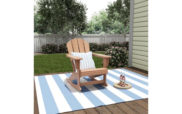 Outdoor brown rocking Adirondack chair in high-density plastic with a transitional style, shown on a blue and white striped patio rug with a striped pillow.
