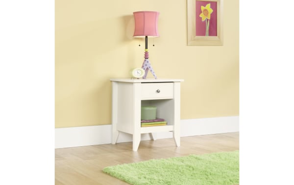 Janis White Nightstand with Drawer in transitional style, made of wood with smooth metal runners, featuring a bottom storage shelf and a pink lamp on top.
