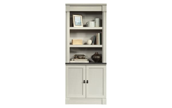 Tall gray wooden bookshelf with two lower cabinet doors, open shelving with decorative items including books, vases, and framed photo, classic design.