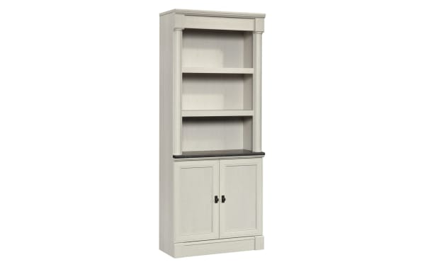 Tall white wooden bookshelf with two open shelves and a two-door cabinet at the bottom, featuring black handles and a dark countertop.