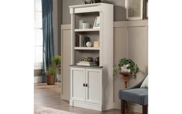 Tall white wooden bookshelf with two lower cabinet doors, open shelving above, decorative molding, and black hardware, styled with books and decor.