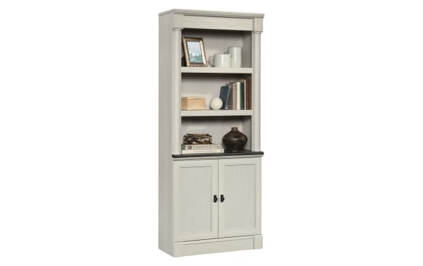 Tall white wooden bookcase with two shelves, decorative items, and a black countertop above double-door storage cabinet.