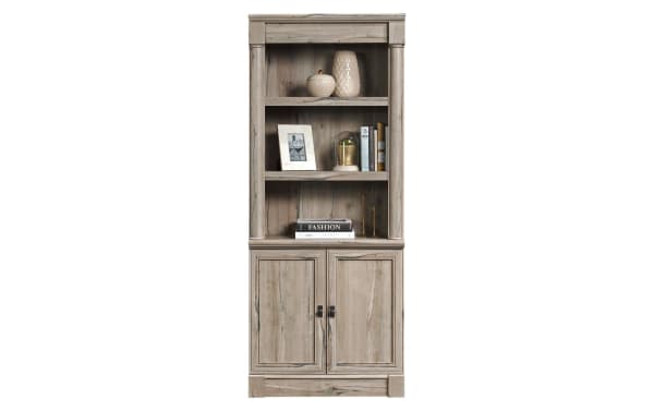 Tall wooden bookshelf and cabinet unit in light weathered gray finish with two open shelves, decorative items, and two-door storage below.
