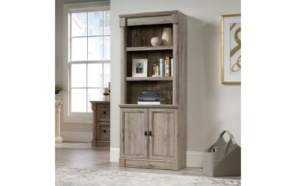 Tall rustic wood bookshelf cabinet with two shelves displaying decorative items and books, and a closed two-door storage compartment below.
