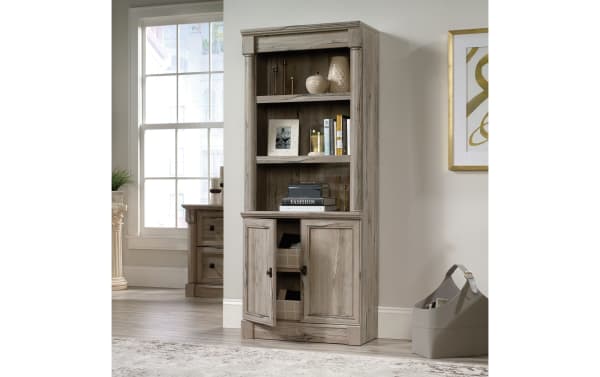 Tall wooden bookshelf cabinet in light gray finish with open shelves and closed storage doors, styled with decor and books, in a modern living room.