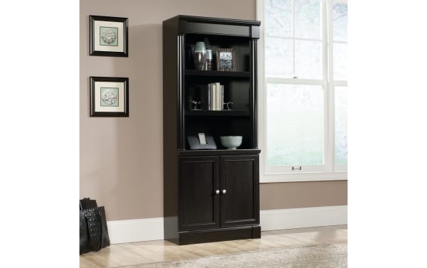 Dark wood bookcase with two shelves, two cabinet doors with silver knobs, decorative columns, and framed art on adjacent wall.