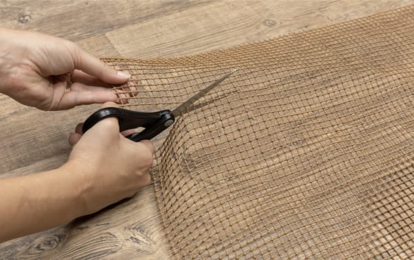 Hands cutting a brown, weather-resistant polyester mesh outdoor rug pad with scissors, designed for slip resistance on stone or wood surfaces.