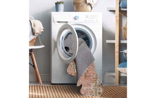 Front-loading washing machine with a pink traditional Kinsley 5' x 7' washable rug made of stain-resistant polyester fabric partially inside, highlighting its machine-washable, non-slip, and durable design for high-traffic areas.