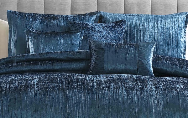 Luxurious dark blue crushed velvet bedding set with multiple textured pillows and a matching comforter on a tufted beige headboard.