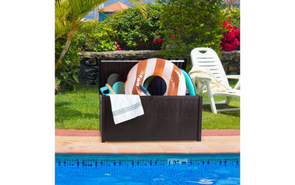 Osgood 113 Gallon Brown Outdoor Storage Box with faux-wood resin slat design, metal frame, and easy-open lid, shown by a pool storing pool toys and towels.