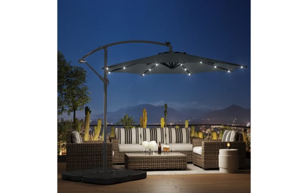 Outdoor patio setup featuring a dark gray cantilever umbrella with built-in LED lights, over a wicker sectional sofa with beige cushions and striped pillows, set against a nighttime cityscape background.