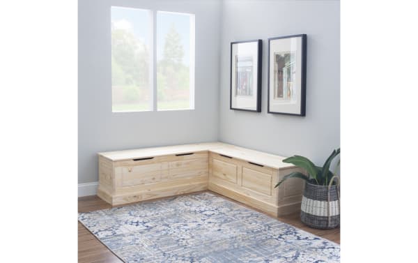 Natural wood L-shaped storage bench with built-in drawers, black cutout handles, placed in a bright corner room with gray walls, framed art, and a blue patterned rug.