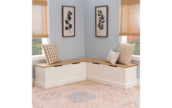 Corner storage bench with white wooden base and natural wood top, featuring beige gingham and striped cushions, set in a light gray room with botanical wall art.