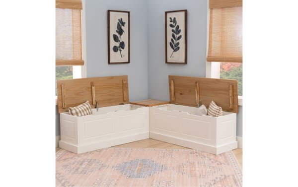 White wooden corner storage benches with hinged natural wood lids open to reveal storage space, placed in a light gray room with framed botanical prints.