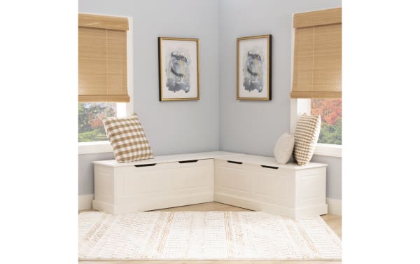 White wooden corner storage bench with paneled design, beige and brown checkered cushions, light gray walls, bamboo blinds, and abstract wall art.