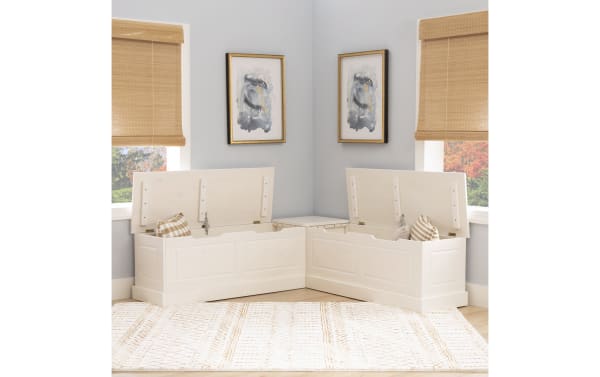 White wooden corner storage bench with hinged lids, beige and white plaid cushions, beige woven window shades, and a patterned beige rug.