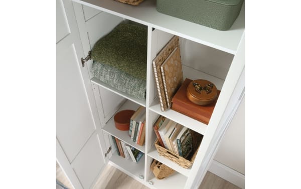 Bella White 2 Door Storage Cabinet in contemporary style, made of metal and particle board, with four adjustable shelves holding blankets, books, and decor.