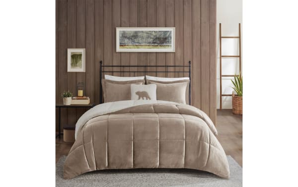 Taupe velvet comforter set on a black metal bed frame with matching pillows, a white accent pillow with bear design, and rustic wood wall background.