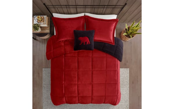 Red velvet comforter set with matching pillow shams and a black throw pillow featuring a red bear design, displayed on a metal bed frame with a wooden nightstand and potted plant.