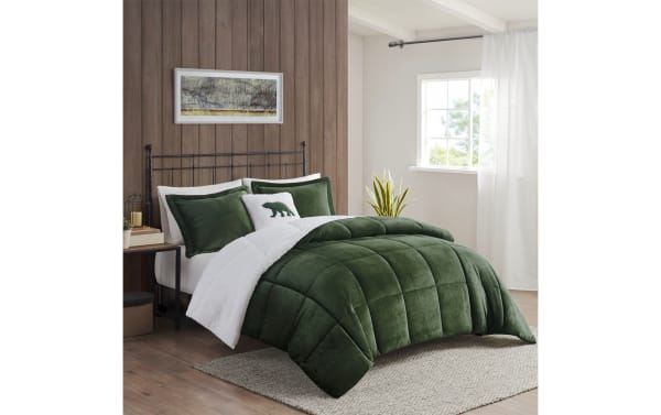 Dark green plush velvet comforter set with matching pillow shams and white sherpa fleece blanket on bed in modern bedroom with wood panel wall and window.