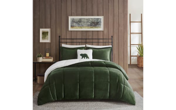 Green velvet comforter set on black metal bed frame with white sherpa lining, decorative bear pillow, wooden wall, and rustic decor.