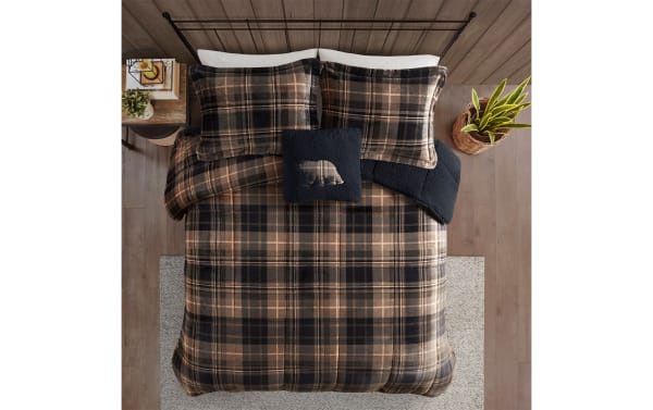 Cozy bed with brown and black plaid comforter set, matching pillowcases, and navy blue bear-themed accent pillow on wooden floor.