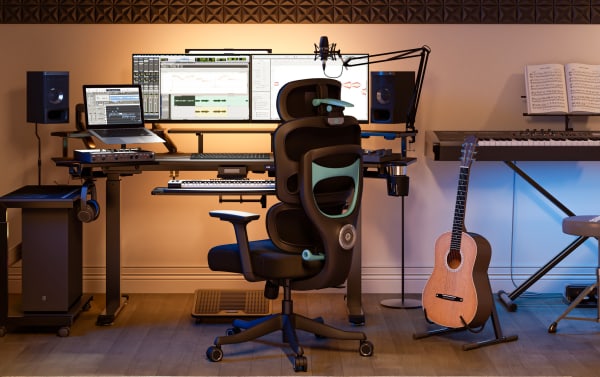 Modern music production workspace with dual monitors showing audio software, ergonomic black office chair, Yamaha keyboard, guitar, and laptop.