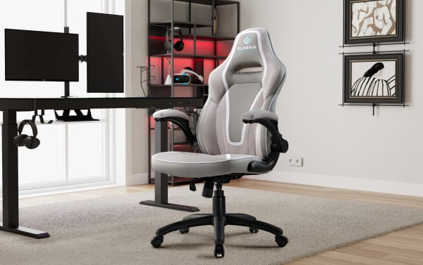 Eureka Ergonomic gray and white padded gaming chair with adjustable armrests and black swivel base, set in a modern office with dual monitors and shelving.