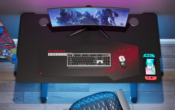 Aurora Black Adjustable Gaming Desk with carbon-fiber textured particle board top, powder-coated metal legs, dual cable holes, socket holder, and height-adjustable bases. Transitional style gaming desk with spacious surface, featuring a curved ultra-wide monitor, mechanical keyboard, gaming mouse on a large Eureka Ergonomic mousepad, and a Nintendo Switch.