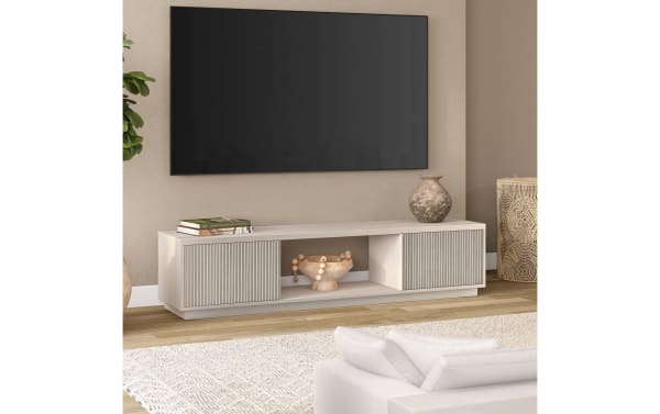 Modern beige wooden TV stand with vertical slat cabinet doors, open central shelf, decorative vase, and books under a wall-mounted flat-screen TV.