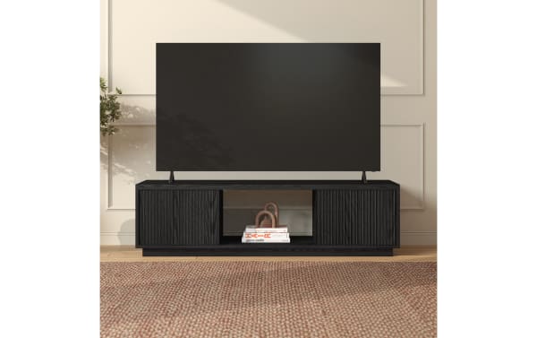 Black wooden TV stand with vertical grooves on cabinet doors, open central shelf holding books, supporting a large flat-screen TV, set against a beige paneled wall.