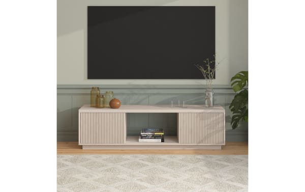 Modern beige wooden TV stand with vertical grooves, open shelf with books, decorative glass jars, and a plant in a minimalist living room setting.