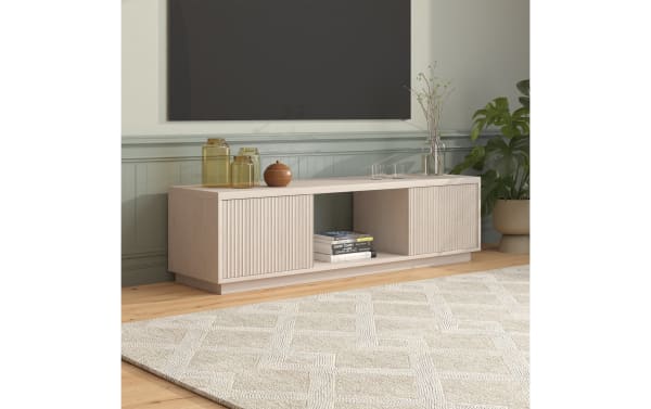 Light wood TV console with vertical slat cabinet doors, open shelf with books, and decorative glass jars and vase, set against a muted green wall.