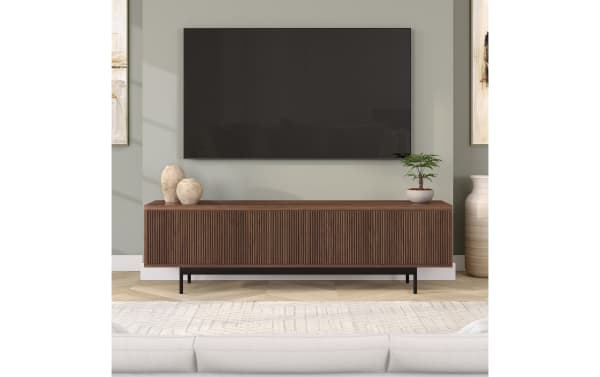 Modern living room setup featuring a wall-mounted flat-screen TV above a mid-century style wooden media console with vertical grooves, dark wood finish, and black metal legs. Decor includes two beige ceramic vases and a small potted plant, set against a light gray wall with framed abstract art partially visible.