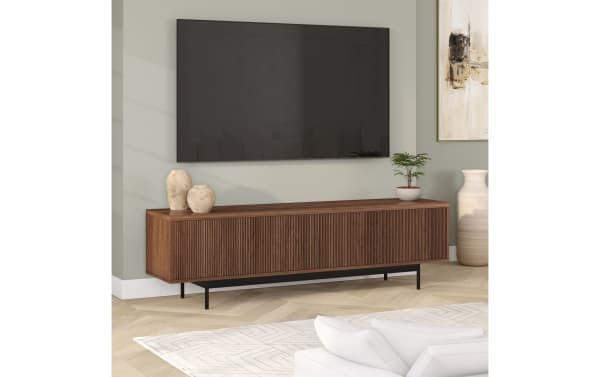 Modern wooden TV console with ribbed front panel and black metal legs, styled with ceramic vases and a small potted plant, under a wall-mounted flat-screen TV.
