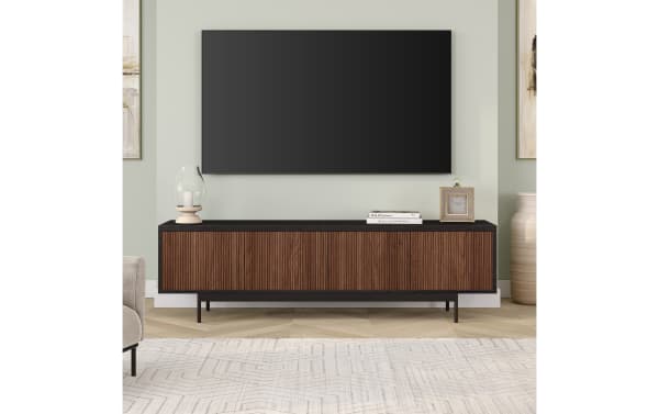 Modern living room setup featuring a wall-mounted flat-screen TV above a black and dark brown wood media console with ribbed front panels, decorated with a glass candle holder, two stacked books, and a small square clock. The room has light green walls, light wood herringbone flooring, and a beige patterned rug.