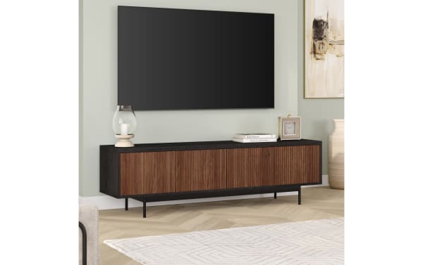 Modern black and brown wood TV stand with ribbed cabinet doors, slim metal legs, and decorative items beneath a wall-mounted flat-screen TV.