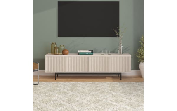 Modern beige wooden TV console with vertical grooves, black metal legs, decorative glass jars, books, and a vase with branches against a green wall.