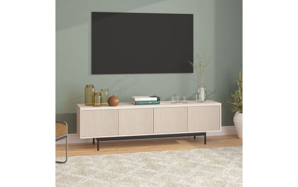 Modern beige wood TV console with vertical grooves, black metal legs, decorative glass jars, books, and a vase with branches against a sage green wall.