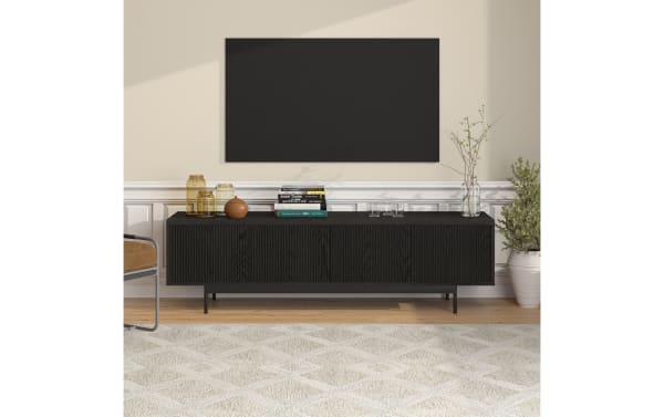 Modern living room setup featuring a black ribbed wood media console with metal legs, decorated with glass jars, books, and plants under a wall-mounted flat-screen TV.