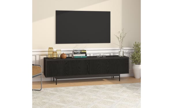 Modern black wood media console with ribbed front doors, metal legs, and decorative jars, books, and plants on top, set in a living room.