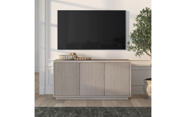 Modern beige wooden sideboard with vertical groove detailing, decorated with a ceramic bowl and wooden beads, under a wall-mounted flat-screen TV, next to a potted olive tree on light hardwood flooring.