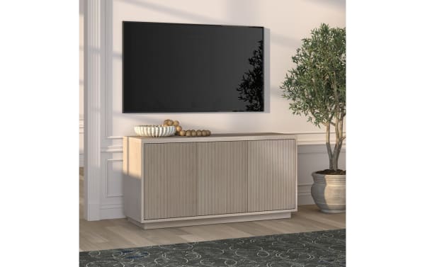Light wood TV console with vertical slat cabinet doors, a decorative bowl, and wooden beads on top, paired with a wall-mounted flat-screen TV and a potted olive tree.